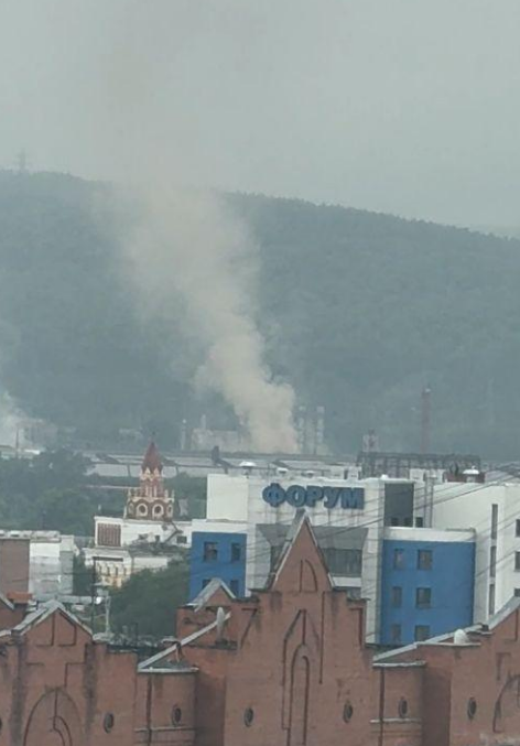Панорама индустриальной зоны Новокузнецка с промышленными трубами, выделяющими плотные клубы серого дыма в атмосферу.